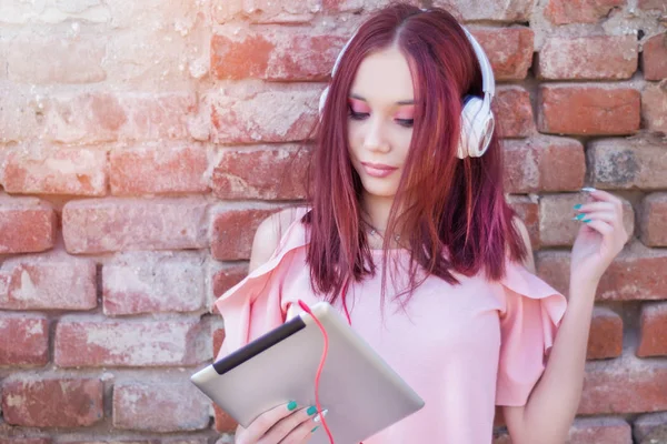 Cute redhead girl listening to music on digital tablet with headphones against brick wall outdoor. Enjoy in life and music concept. Space for text. Close up, selective focus