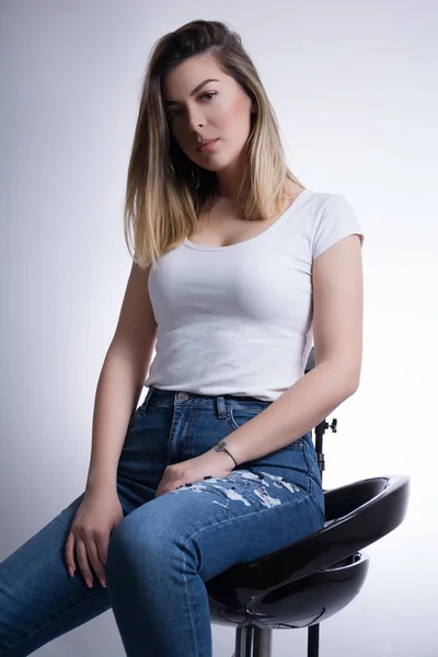 Female in white t shirt and in blue jeans sits on a bar stool and posing in studio. Woman portrait on white background and fashion concept. Close up