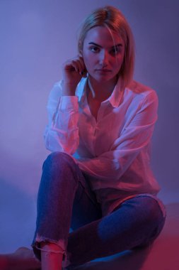 Renkli arka planda seksi genç kadın portre. Mavi kot baştan çıkarıcı sarışın model kız. Kız oturuyor. Kırmızı ve mavi ışık. Gece hayatı