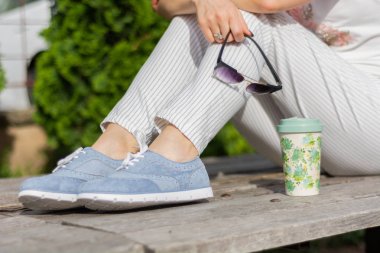 Çizgili pantolon ve mavi spor ayakkabı ile modern bir kadın parkta bir bankta oturan ve güneş gözlüğü tutan, kız bacaklarının yanında kahve fincanı. Kadınlık moda konsepti. Yakın çekim, seçici odak