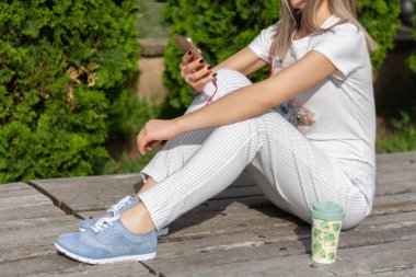 Kadın akıllı telefonda çalışıyor ve güneşli bir bahar gününde parkta bir fincan kahvenin yanında bankta oturuyor. Çizgili pantolonlu ve mavi ayakkabılı şık bir kadın. Yaklaş, seçici odaklanma