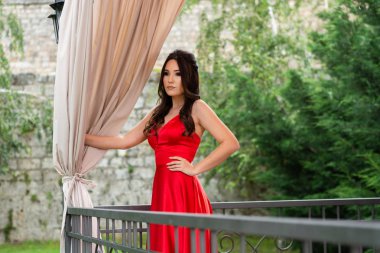 Kırmızı elbiseli kız poz ve bir yaz günü restoran bahçesinde balkon ve perdeler çit arkasında kalça bir el tutar. Moda ve güzellik konsepti. Yakın çekim, seçici odak