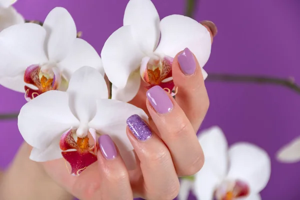 Young girl hand with a lilac color nails polish gel and beautiful orchid flower decoration on purple background in the studio. Manicure and beauty concept. Close up, selective focus
