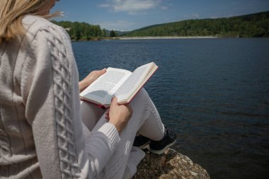 Güneşli bir günde göl kenarında kazakla kitap okuyan bir kadın. Gölün kıyısındaki büyük bir kayanın üzerinde bacaklarla ilgili bir kitapla oturan bir kız. Rahatlama konsepti. Yaklaş, seçici odaklanma