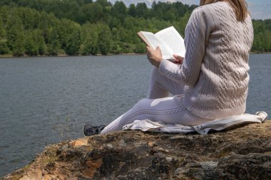 Doğada bir gölün kenarında oturan ve kitap okuyan bir kadın. Gölün kıyısındaki büyük bir kayanın üzerinde oturan kazaklı sarışın kız. Rahatlama konsepti. Yaklaş, seçici odaklanma