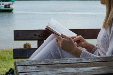 Genç kız bankta kitap okuyor ve arka planda göl suyu var. Tahta bir bankta oturan sarışın kadın, elinde bir kitapla. Rahatlama ve eğitim kavramı. Yaklaş, seçici odaklanma