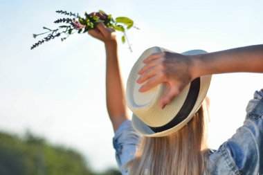 Elinde çiçeklerle havaya yükselen ve kafasında beyaz bir şapka tutan genç bir kız. Güneşli bir yaz gününde sarışın, mutlu bir kız. Doğa konseptinde rahatlama. Yaklaş, seçici odaklanma