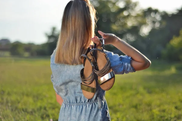 Girl holding summer sandals on back in nature on summer sunny day. Blonde woman enjoying and walking outdoor in the park on good weather. Relaxation in nature concept. Close up, selective focus