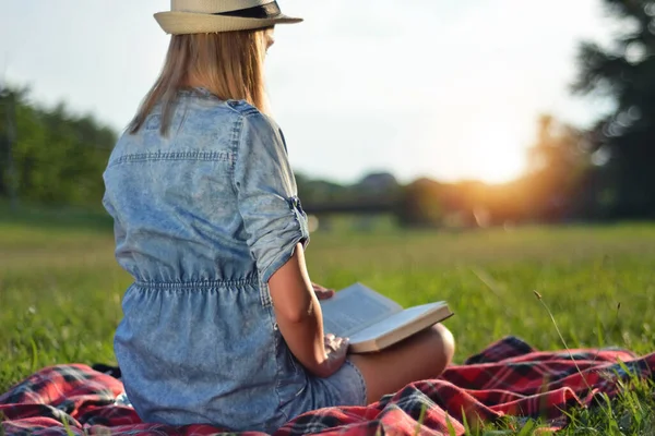 Woman with hat reading book in nature on summer sunset day in the park. Girl sitting on plaid blanket in meadow and sunshine in background. Relaxation in nature concept. Close up, selective focus