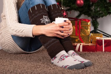 Elinde bir fincan kahve tutan ve halı döşemesinde oturan genç kız, Noel ağacı ve arka plandaki hediye kutusu, kapat.