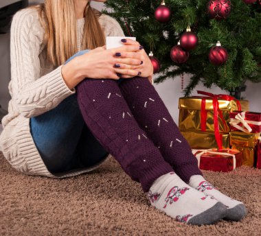 Genç kız yerde oturuyor ve elinde bir fincan kahveyle ayaklarının üzerinde duruyor. Arka planda ısıtıcılar ve Noel ağacı var. Yakın plan.