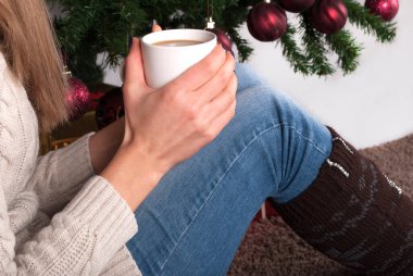 Kız ve kız bacaklarına bir fincan kahve. Arka planda ısıtıcılar ve Noel ağacı var. Kapatın.