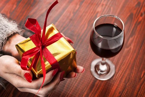 Female hand holding golden box present with red bow on wooden background and wine glass on table