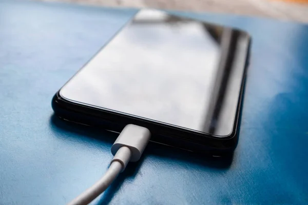 White phone charging cable plugged into a smartphone on a blue desk at home. Smart technology and charging concept. Copy space for design, close up, selective focus