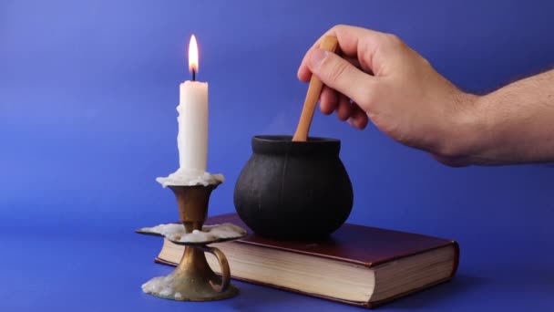 Чорний відьомський горщик курить на старій книзі та руці, змішуючи з дерев'яною ложкою та свічкою під час спалювання свічки — стокове відео