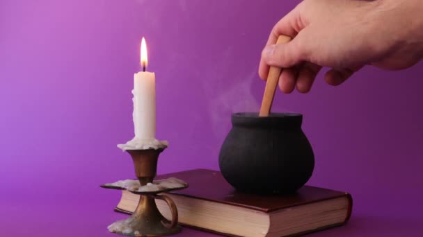 Olla bruja negra está fumando en el libro viejo y la mano mezclando con una cuchara de madera, y una vela en candelabro — Vídeos de Stock