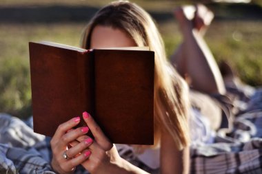 Yaz günü gün batımında parkta kitap okuyan bir kız. Kadın, battaniyenin üzerine uzanmış ve elinde açık bir kitap tutuyordu. Doğa konseptinde rahatlama