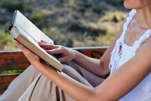Woman holding an open book in hands and reading. Close up of girl sitting on a bench in the park on a summer day
