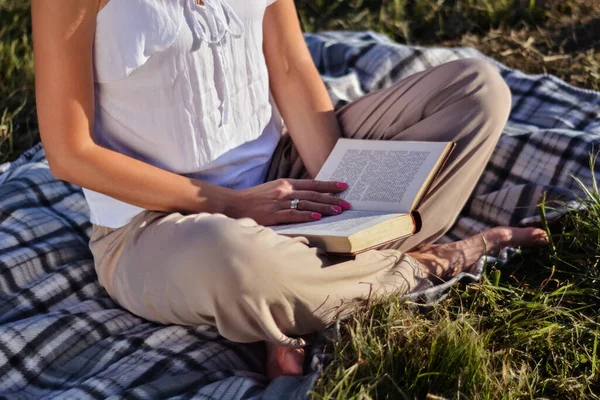 Genç bir kadın doğada kitap okuyor ve sıcak yaz günbatımında battaniyeye oturuyor. Rahatlama ve zevk konsepti. Yaklaş, seçici odaklanma