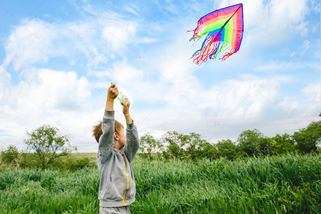 Cute white kid playing colorful kite outdoor at summer meadow or park. Color photography of caucasian young boy isolated at sunny blue sky and green field background.