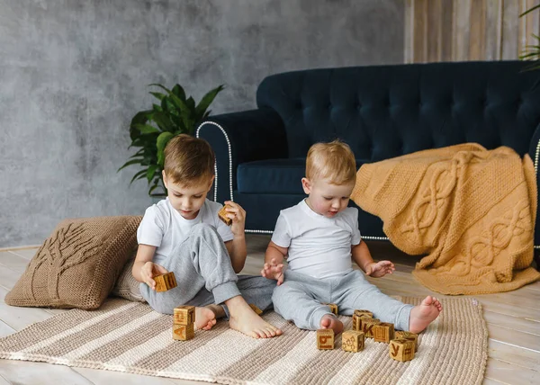 Two boys brothers plaing with wooden cube blocks on knitted carpet on the floor indoor. Knitted style in the interior: pillows, plaid and carpet. Warm and cozy.