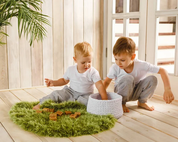 Due fratelli ragazzi che intrecciano con blocchi di cubo di legno su tappeto di erba lavorato a maglia sul pavimento al coperto. Stile lavorato a maglia all'interno: tappeto e cestino. Caldo e accogliente. Bambini giocosi — Foto Stock