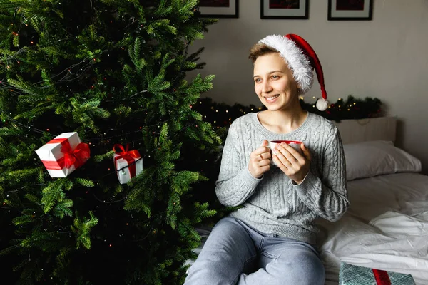 Kapalı mekanda sıcak noel aktivitesi: kırmızı Noel Baba şapkalı kadınlar gülümsüyor, bir bardak çayı noel ağacının yanında tutuyorlar. Tatilin beklentisi.