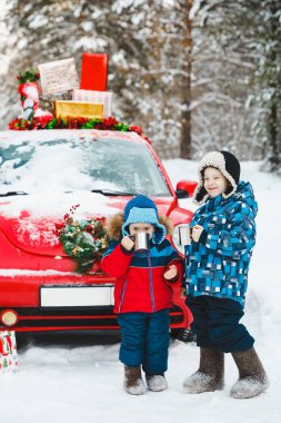 Noel açık hava aktivitesi: iki çocuk kışın karlı ormanda içinde hediyeler olan kırmızı bir arabanın yanında çay içerler. Yeni Yıl