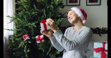 Kapalı alanda Noel Baba şapkalı kadınlar gülümser, hediye kutusunu Noel ağacının yanında tutarlar. Kız süslü hediyeye hayran. Tatilin beklentisi.