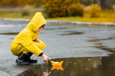 Sarı su geçirmez pelerinli ve lastik çizmeli çocuk sonbaharda yağmurda açık havada kağıt el yapımı tekne oyuncağıyla oynuyor..