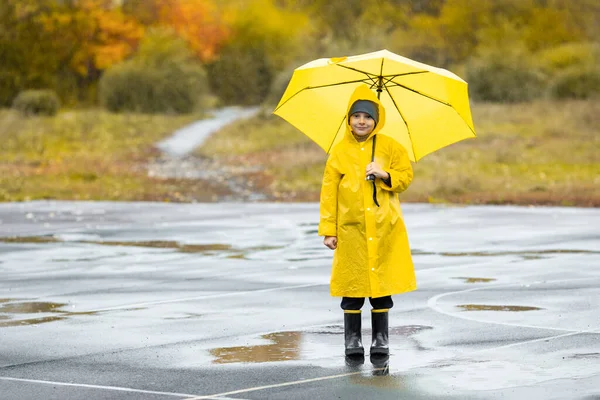 Sarı su geçirmez pelerinli ve lastik botlu çocuk sonbaharda yağmurda açık havada su birikintisinde duruyor.. — Stok fotoğraf