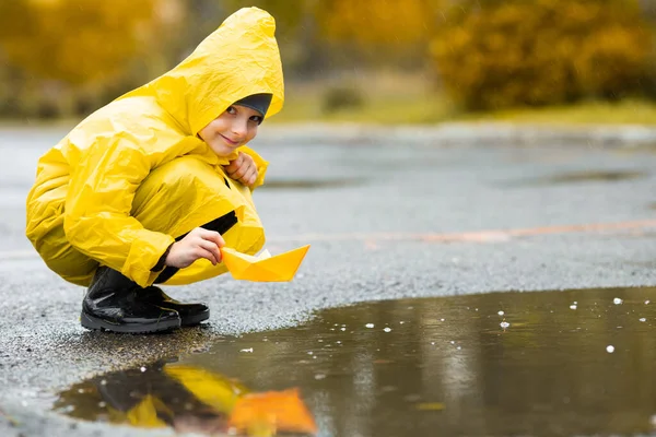 Sarı su geçirmez pelerinli ve lastik çizmeli çocuk sonbaharda yağmurda açık havada kağıt el yapımı tekne oyuncağıyla oynuyor.. — Stok fotoğraf