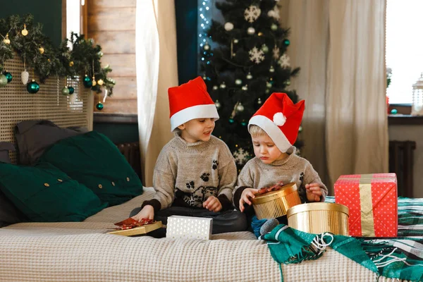 Actividad navideña en interior: dos chicos con sombreros de Santa Claus sostienen y consideran cajas con regalos, sentados en una cama cerca de un árbol de Navidad con luces. Anticipación de las vacaciones. —  Fotos de Stock