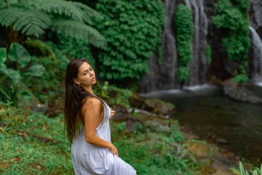 Beyaz elbiseli güzel bir kız kayalıklarda yürür, bir su şelalesinin önünde.