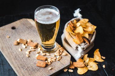 Bir bardak taze bira, patates cipsi ve siyah arka planda fıstık.