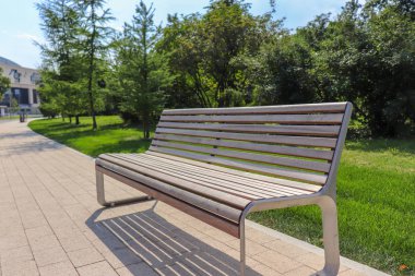 Bir ahşap Bank Şehir Parkı içinde kalan için. Yeşil iğne yapraklı ağaçlar içinde belgili tanımlık geçmiş. Taş yol yayalar için. Yeşil çim çim.