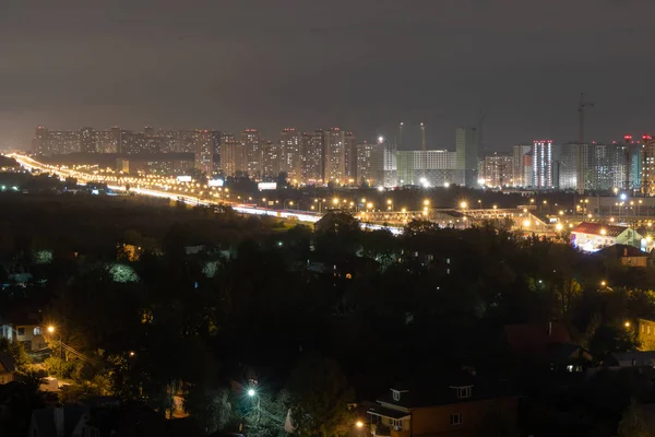 Evening city lights are lit from the windows. Road with a passing vehicle. Construction cranes at night. Dark evening sky, burning street lamps on the pillars.