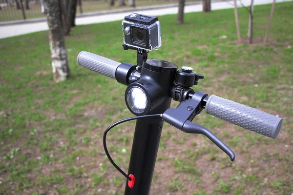 Scooter'ın tekerleği yakın çekim, bir aksiyon kamerası takılı. Gri kulplu elektrikli scooter siyah.