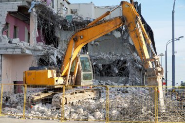 Sarı ekskavatör bir araç ile çok katlı bir bina yıkar. Binanın yıkılmış zeminleri, taş parçaları, beton, bağlantı parçalarıdır. Çelik çit çit.