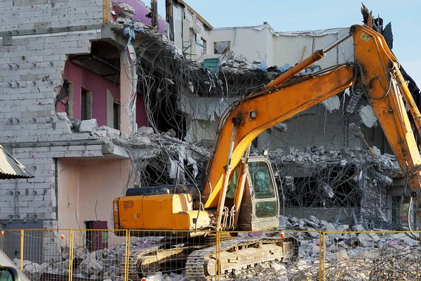 Sarı ekskavatör çok katlı bir binayı yıkıyor. Binanın yıkılmış zeminleri, taş parçaları, beton, bağlantı parçalarıdır. Taş ve armatürler üzerinde inşaat ekipmanları. 