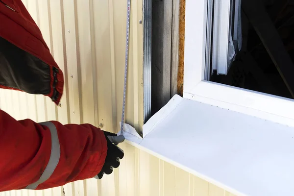 Um construtor de luvas mede a janela do lado de fora com uma fita métrica. Profissional artesão em um manto vermelho com uma ferramenta de medidas ao ar livre . — Fotografia de Stock