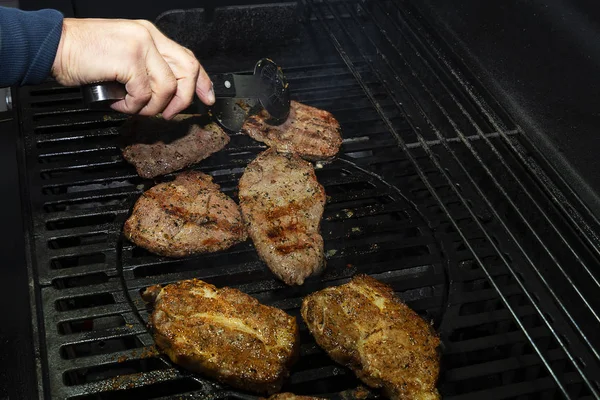 Vários pedaços suculentos de close-up de carne são cozidos em um churrasco. A carne marinada com especiarias é grelhada em uma grelha a gás de ferro. Mão vira pedaços de carne de porco . — Fotografia de Stock