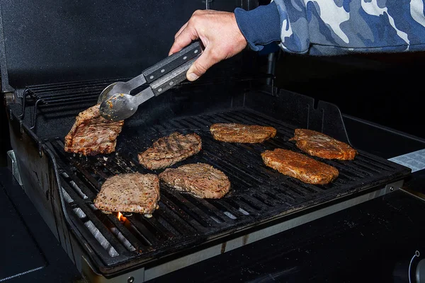 Ręka odwraca kawałek soczystego mięsa z grilla. Grill gazowy jest czarny, gotuje pyszne mięso. — Zdjęcie stockowe