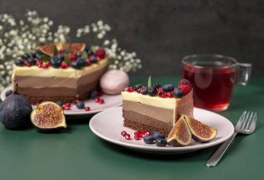 Pembe tabakta bir parça kek, yanında taze böğürtlen ve koyu yeşil arka planda bir kupa çay.