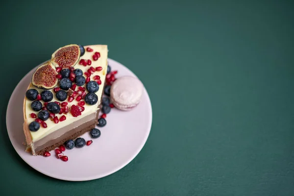 Pembe tabakta bir parça kek, taze böğürtlen ve badem kurabiyesi koyu yeşil arka planda bir fincan çay.