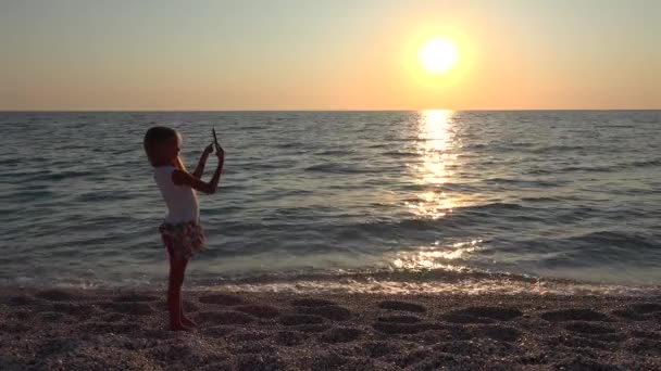 女孩在日落时通过智能手机在海滩上自拍孩子玩平板电脑 — 图库视频影像