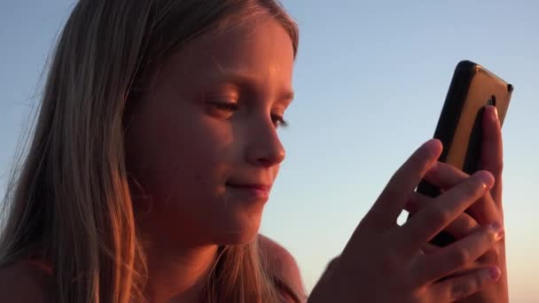 Copil care joacă smartphone, copil pe plajă la apus de soare, fată care folosește tabletă pe malul mării — Videoclip de stoc