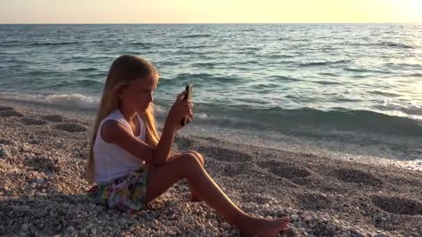 Dziecko bawiące się na smartfonie, Dziecko na plaży o zachodzie słońca, Dziewczyna korzystająca z tabletu na wybrzeżu — Wideo stockowe