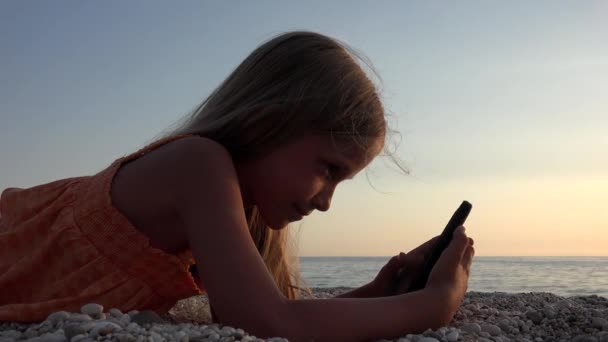 Ребенок играет в смартфон, ребенок на пляже на закате, девушка с планшета на берегу моря — стоковое видео