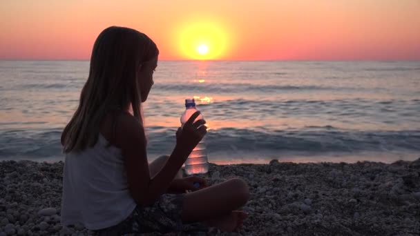 Criança bebendo água na praia ao pôr do sol, Criança assistindo ondas do mar na costa, Menina no litoral — Vídeo de Stock